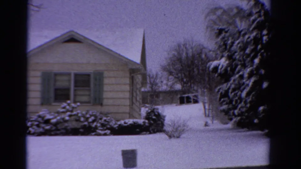 1958 Ontario 8mm frame showing a snow-covered single-story suburban house, green shutters, shrubs, bare trees, and a distant steeple.