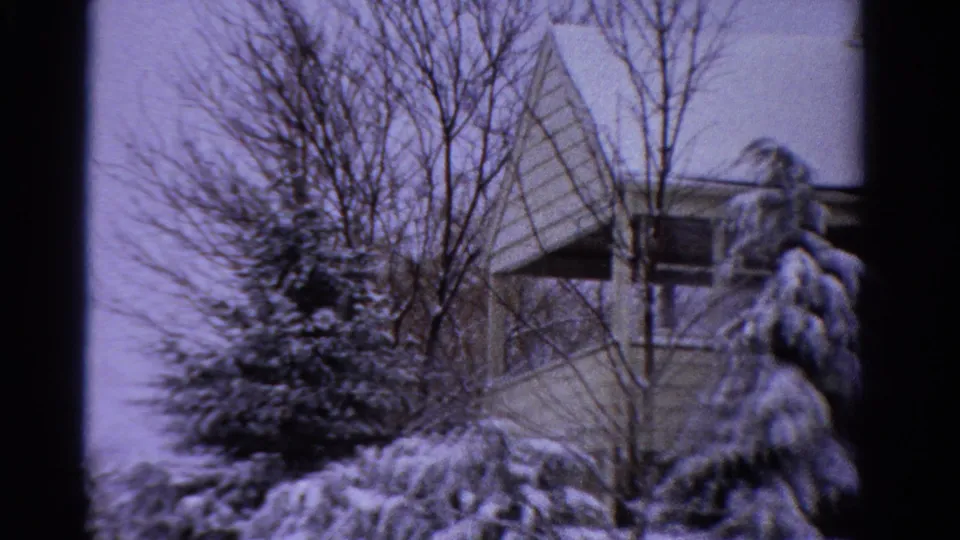 1958 Ontario home-movie frame showing snow-laden evergreens, bare branches, and part of a suburban house roofline.