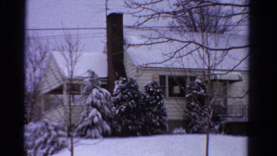 Wider 1958 Ontario view of a snow-covered yard, brick chimney, pale house exterior, evergreen shrubs, and bare winter trees.