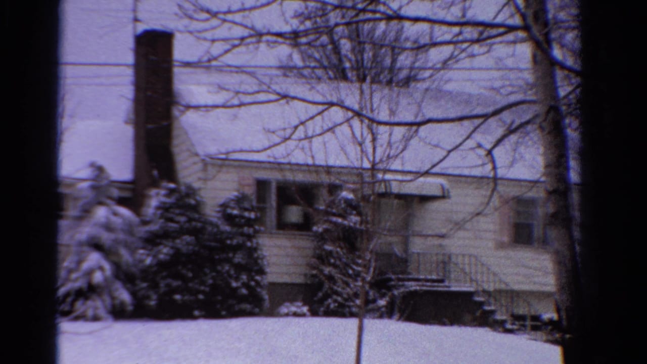 1958 Ontario home-movie frame of a snowy house exterior with a brick chimney, bare trees, shrubs, and pale winter sky.