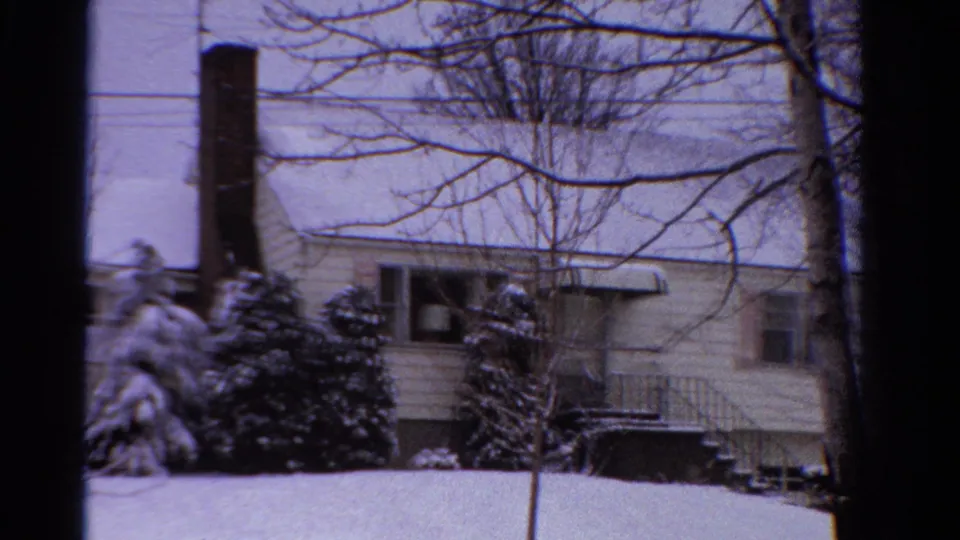 1958 Ontario home-movie frame of a snowy house exterior with a brick chimney, bare trees, shrubs, and pale winter sky.