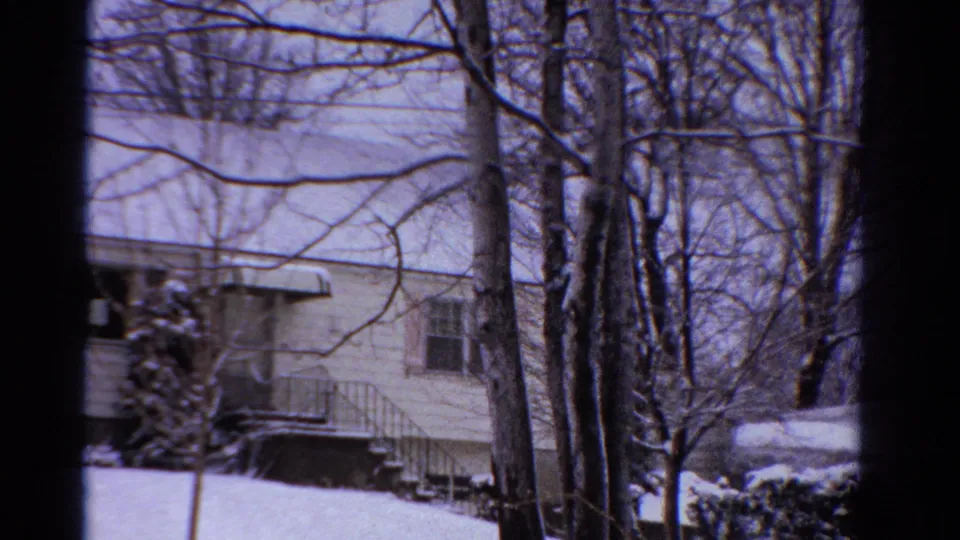 1958 Ontario home-movie view of the house corner with snowy steps, metal railing, shrubs, and leafless trees in the yard.