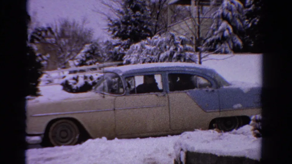 1958 Ontario 8mm frame showing the side of a classic sedan partly surrounded by snow, with an occupant silhouette in the window.