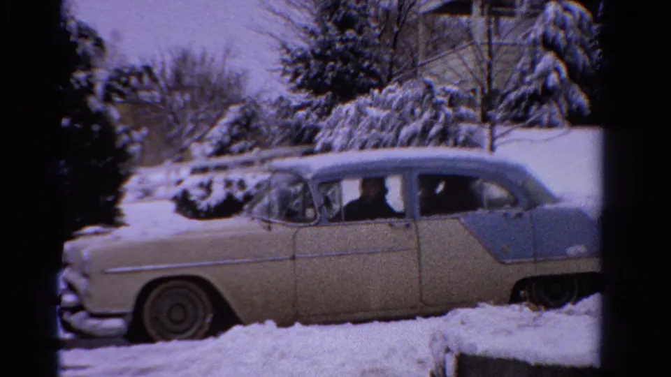 1958 Ontario winter frame showing the two-tone sedan in deep snow, framed by snowy evergreens and a nearby house.