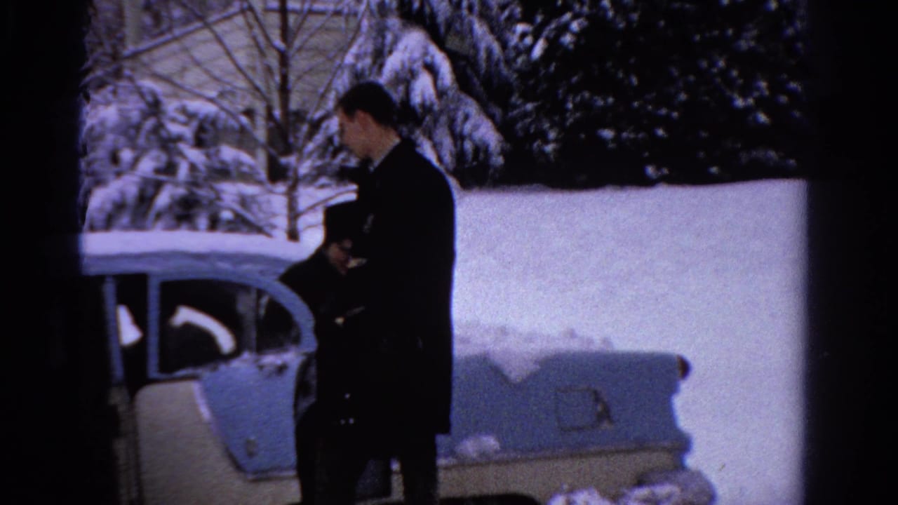 In 1958 Ontario home-movie footage, a man stands near a cream-and-blue sedan parked in deep snow.