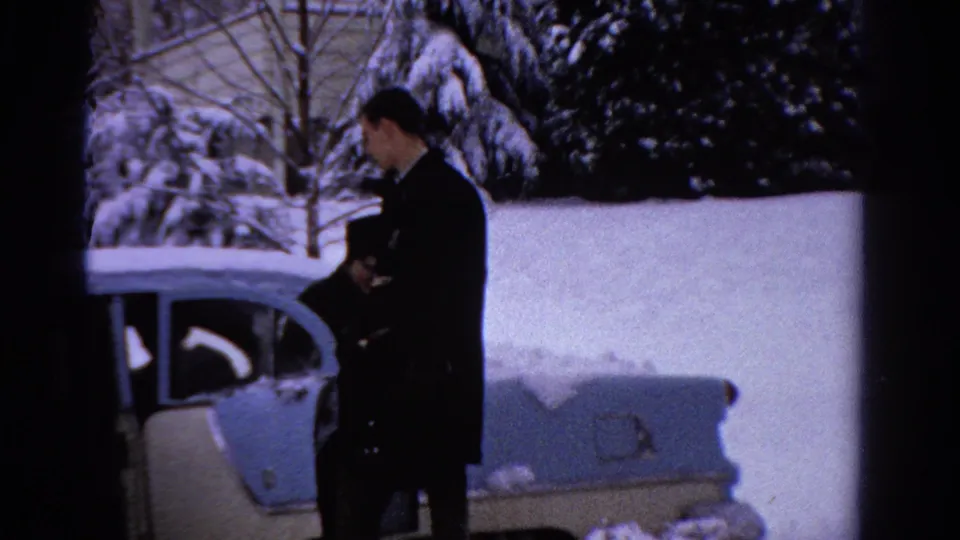In 1958 Ontario home-movie footage, a man stands near a cream-and-blue sedan parked in deep snow.