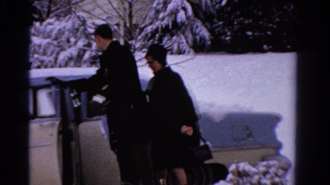 Man opens the rear passenger door as a woman stands near the snowy sedan.