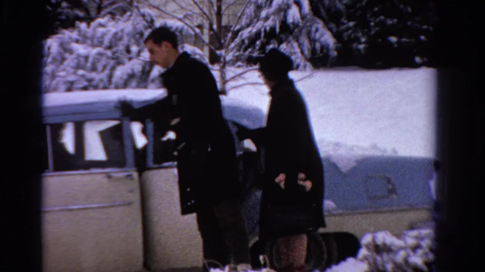 The man reaches toward the sedan door as the woman stands just behind him, framed by snow and evergreen trees.