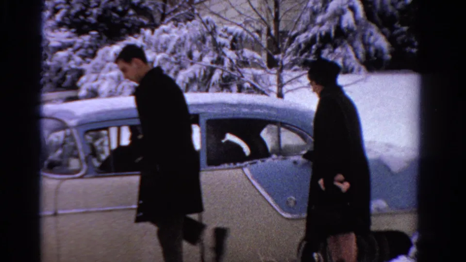 A man and woman in dark winter clothing stand beside the snow-covered two-tone sedan before walking away.