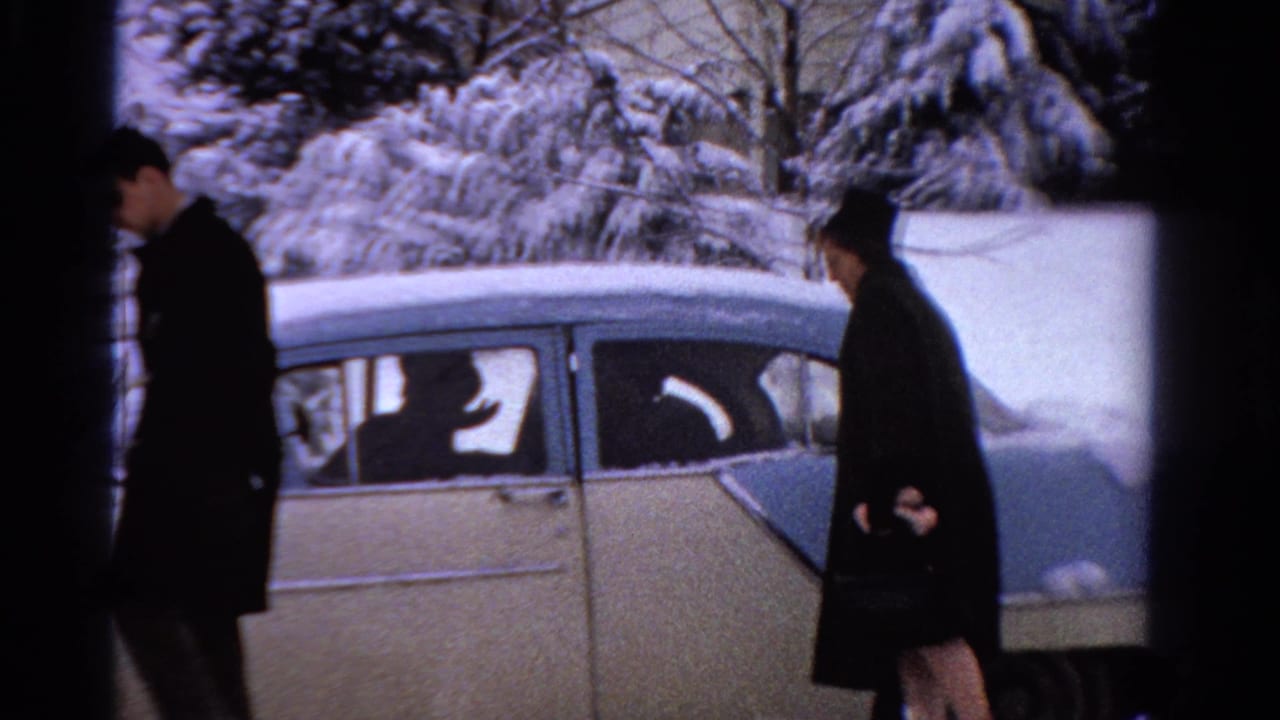 In a 1958 Ontario home-movie frame, two adults in winter coats stand by a snow-covered light-colored sedan.