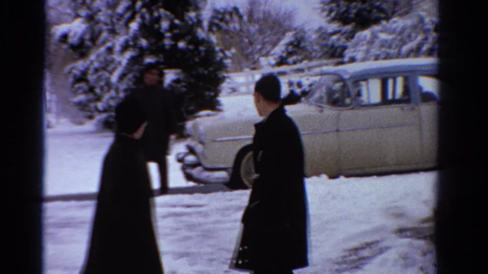 Two adults in dark overcoats stand near the front of a light-colored vintage sedan in snowy 1958 Ontario footage.