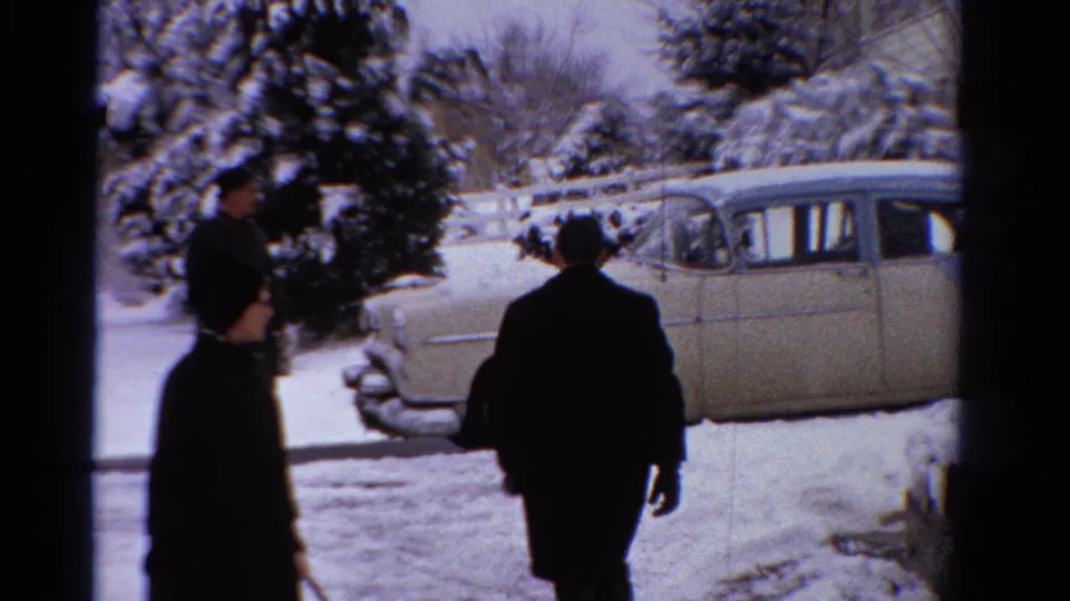 An adult in a dark coat walks toward or past a parked vintage sedan on a snowy Ontario day, with another figure partly visible.