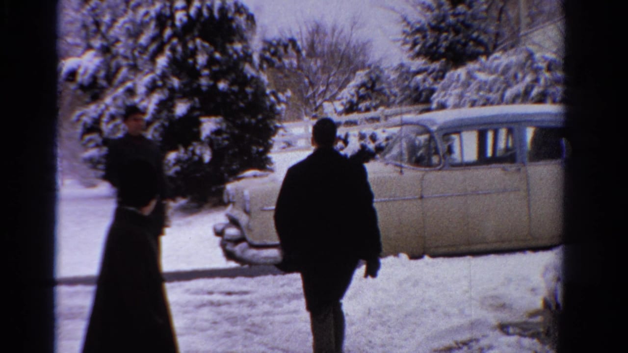 Adults in winter coats walk through snow near a pale mid-century sedan and snowy evergreens.