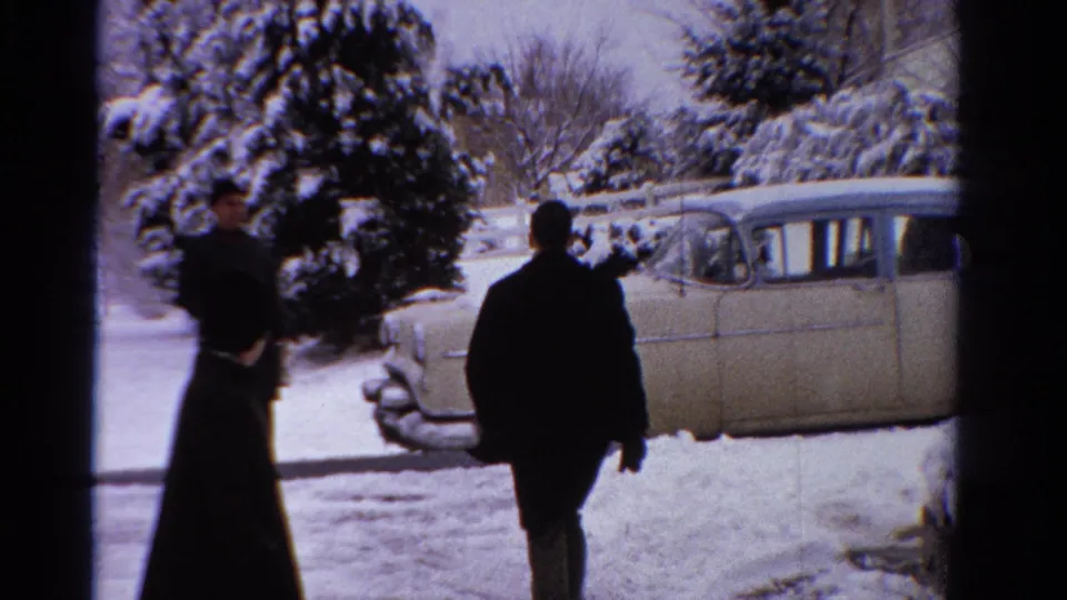 In 1958 Ontario home-movie footage, adults in dark winter coats move across a snowy yard near a pale sedan and snow-laden evergreens.