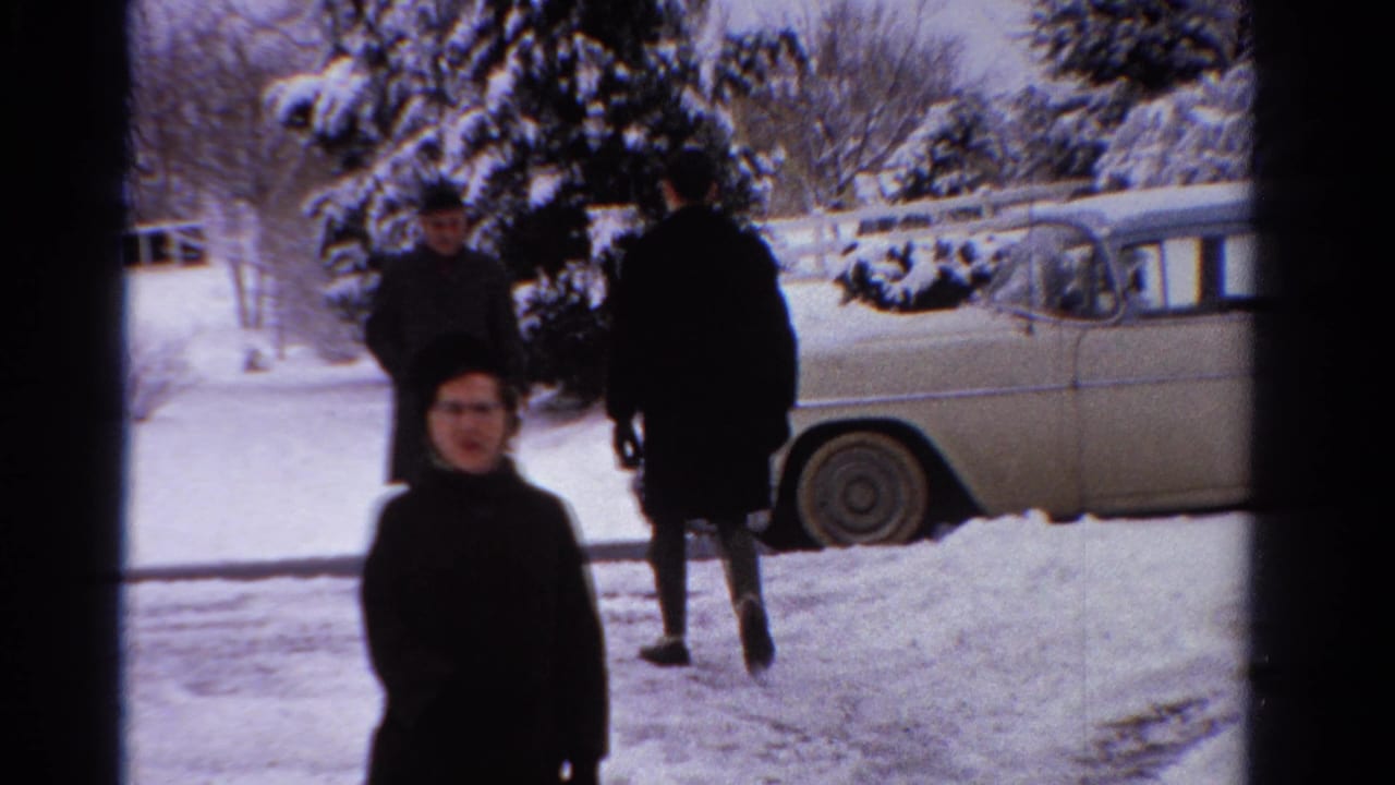 A woman in a dark winter coat faces the camera while two adults move through the snowy Ontario yard near a pale sedan.