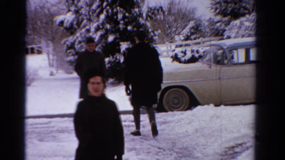 A woman in a dark winter coat faces the camera while two adults move through the snowy Ontario yard near a pale sedan.