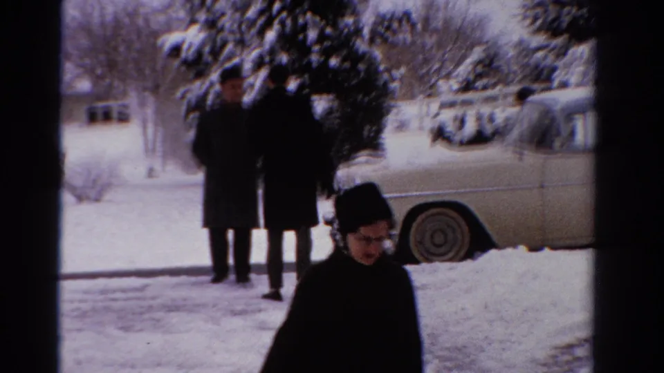 Two adults in long winter coats stand near a snow-covered evergreen and pale sedan as another person crosses the foreground.