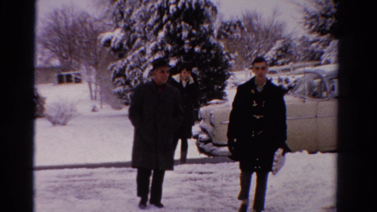 Two men walk toward the camera while a woman follows near a snow-covered car.