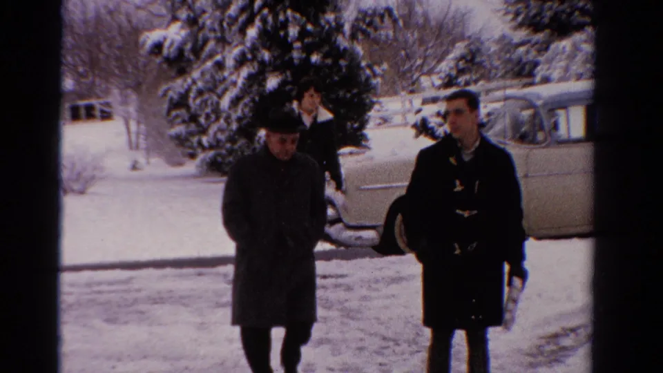 A brimmed-hat man and younger man in a duffel-style coat walk closer to the camera in a snowy 1958 Ontario yard.
