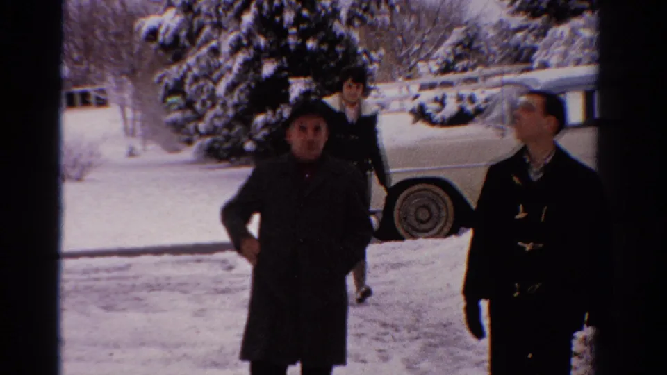 Two men in winter coats pass near the camera in 1958 Ontario, with a woman, snow, and a pale sedan in the background.