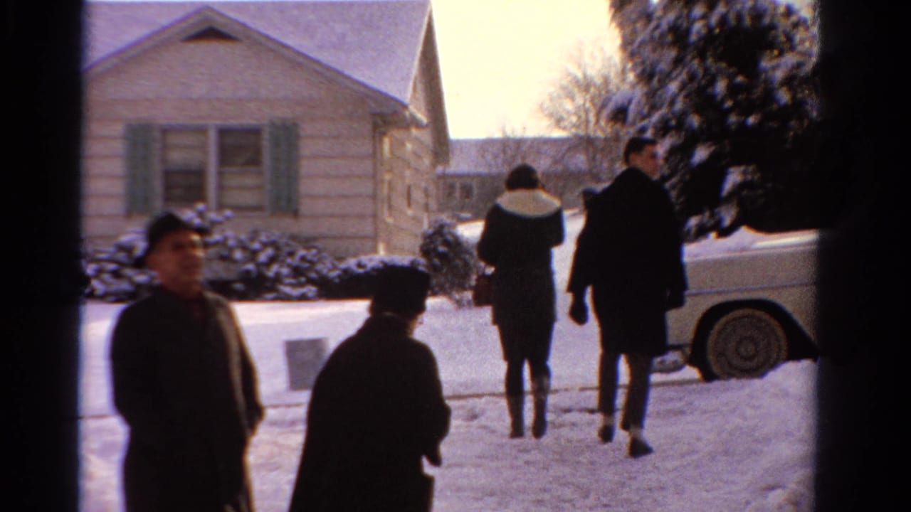1958 Ontario home-movie frame showing adults in winter coats outside a snow-covered house, with a light-colored car parked nearby.