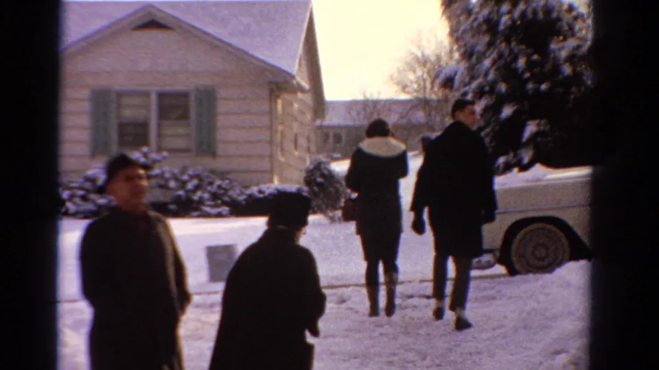 1958 Ontario home-movie frame showing adults in winter coats outside a snow-covered house, with a light-colored car parked nearby.