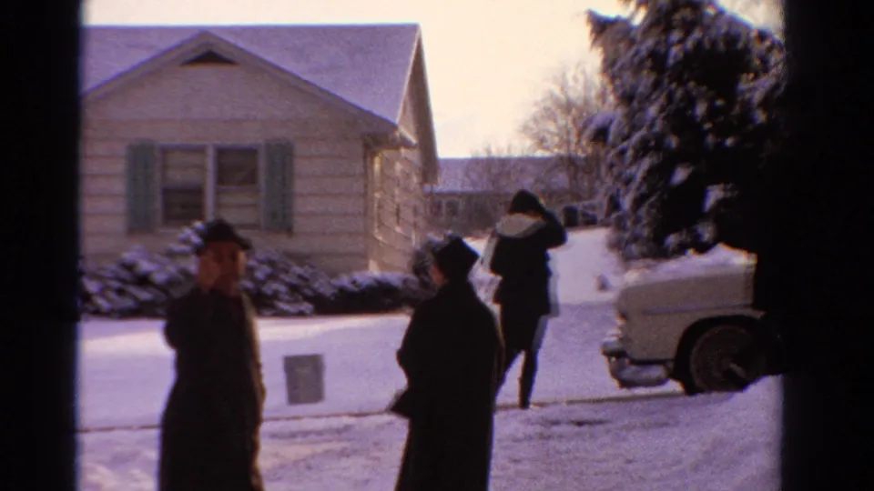1958 Ontario 8mm frame of adults leaving a snowy suburban house, with a light-colored car beside the walkway.