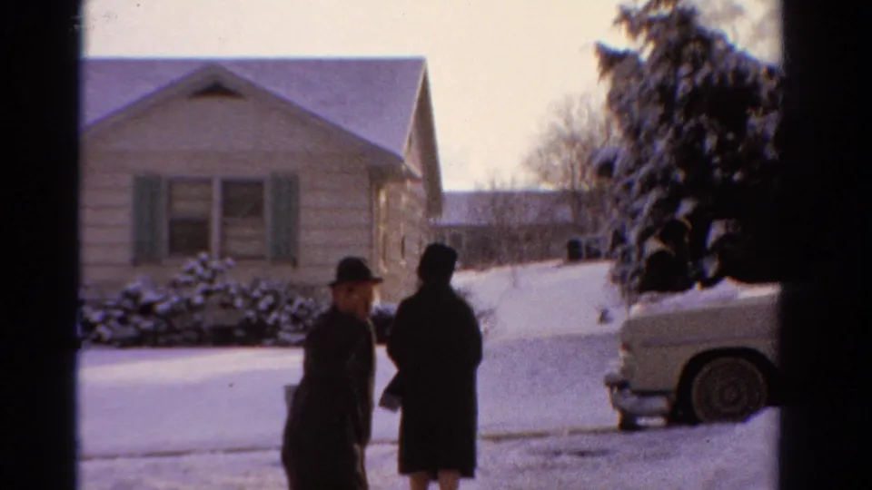 1958 Ontario home-movie frame with adults in dark winter coats, a snowy suburban house, and a light-colored car at the curb.