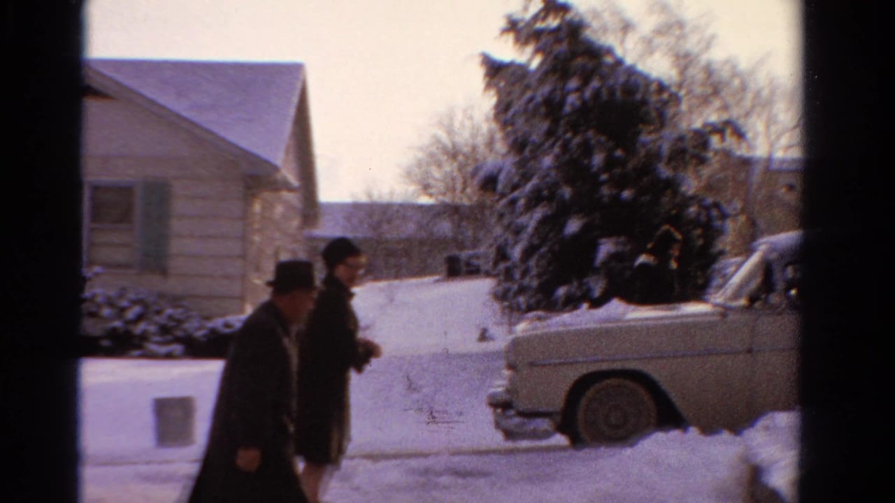 In a 1958 Ontario home-movie frame, two adults in dark winter clothing pass a pale sedan beside snowbanks and a modest house.