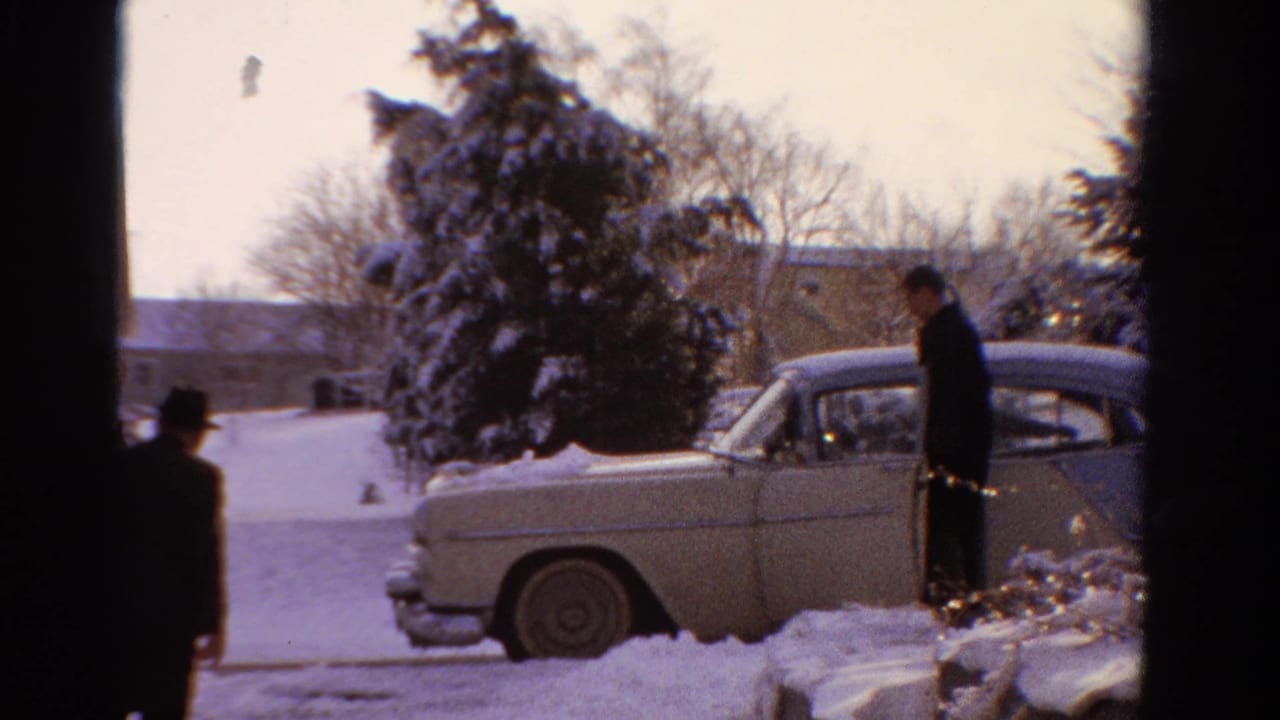 A man stands beside the rear of a pale sedan on a snowy driveway or curb.