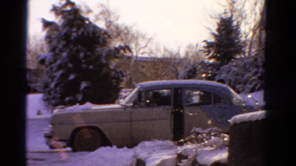The pale two-tone sedan is parked in deep snow with an open door, framed by evergreens and residential houses in Ontario home-movie footage.