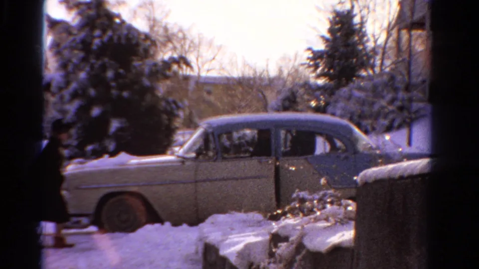 A person in dark winter clothing stands near the open door of a pale sedan in a snowy 1958 Ontario residential scene.