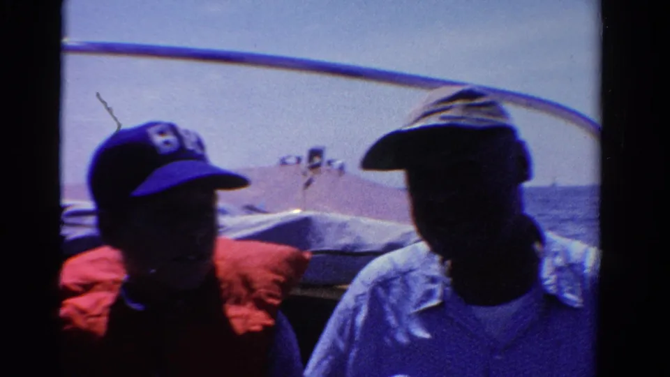 1958 Ontario 8mm frame showing two shadowed passengers seated in a small boat on blue water.