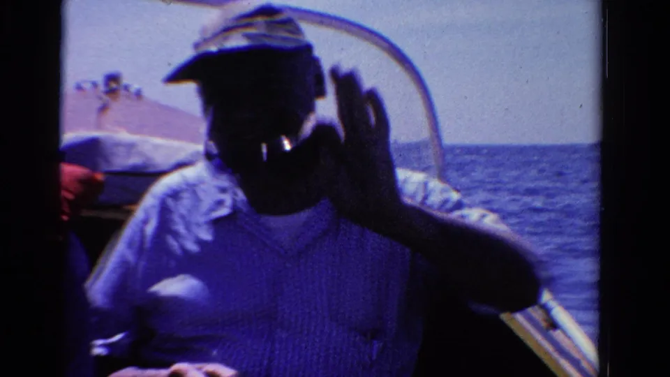 1958 Ontario home-movie frame of a man in a cap waving from a boat, with open water behind him.