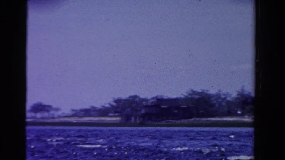 1958 Ontario 8mm frame showing a distant shoreline, trees, a low building, and rippled water.