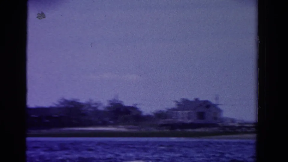 1958 Ontario home-movie frame with a distant waterfront house, trees, shoreline, and blue water.