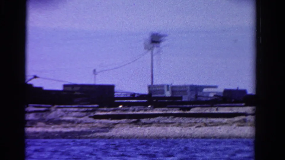 1958 Ontario 8mm frame with low shoreline buildings, a utility pole, and water in the foreground.