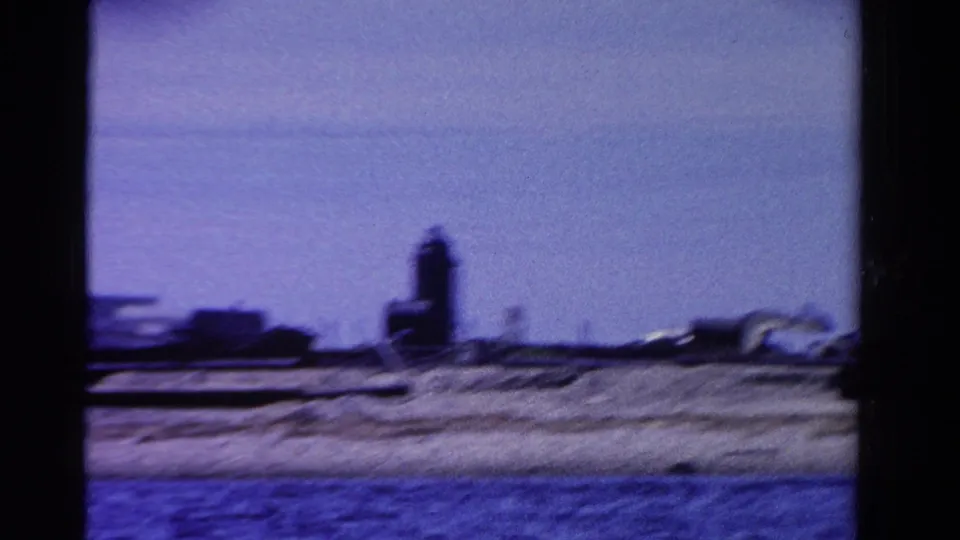 1958 Ontario shoreline frame showing a tall dark tower or tank near low structures, with water in the foreground.