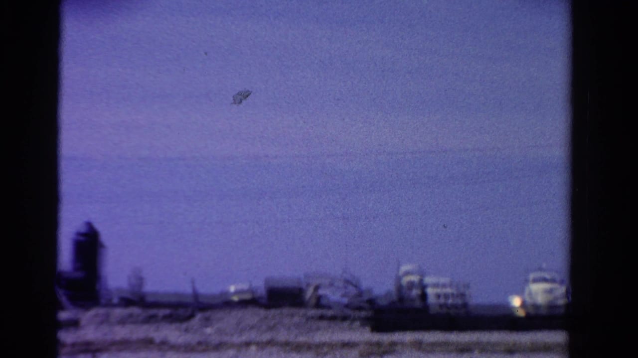 1958 Ontario home-movie frame with distant low buildings, open blue sky, and dark film-edge framing along the sides.