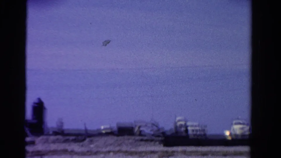 1958 Ontario home-movie frame with distant low buildings, open blue sky, and dark film-edge framing along the sides.