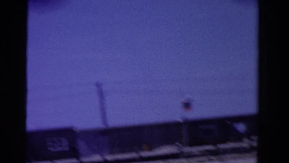 1958 Ontario home-movie frame with low rooftops, a vertical pole-like structure, and a small round roadside fixture under blue sky.