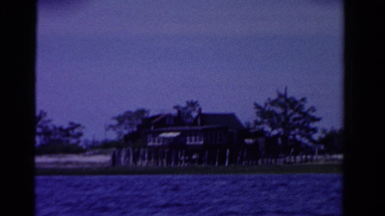 1958 Ontario 8mm frame showing a dark waterfront home, possible dock posts, trees, beach edge, and blue water from a boat.