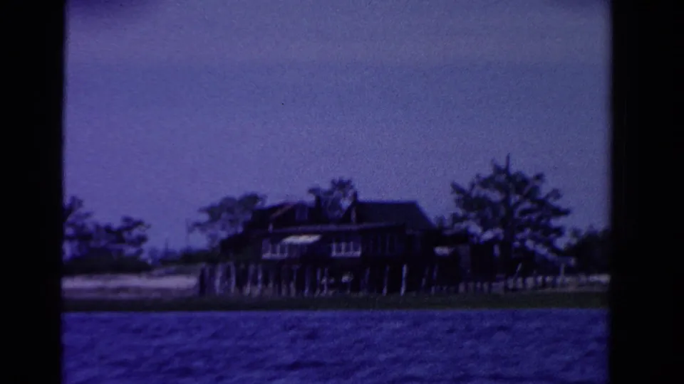 1958 Ontario 8mm frame showing a dark waterfront home, possible dock posts, trees, beach edge, and blue water from a boat.