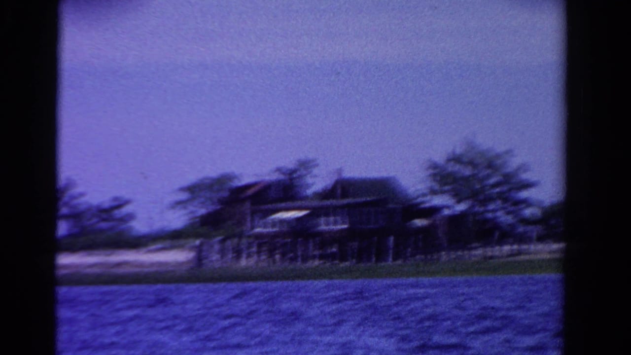 1958 Ontario home-movie frame with a lakeside or coastal home, trees, pale shoreline, and water blurred by boat movement.
