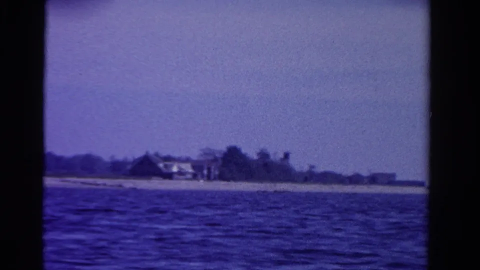 1958 Ontario 8mm view of distant waterfront buildings, a sandy beach, tree line, and calm water from offshore.
