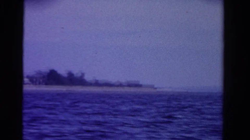 1958 Ontario frame showing rippled water, a low sandy shoreline, wooded trees, and a hazy blue sky from a boat.