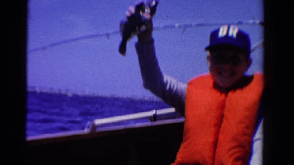 Adult man on an Ontario boat in 1958 holds up a small fish while wearing an orange life vest and blue cap.