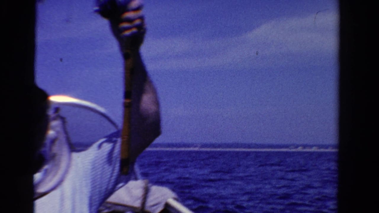 Man on a boat holds a fishing rod or line over blue water under bright sun.
