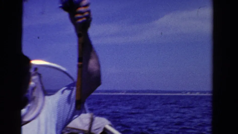 1958 Ontario home-movie frame showing an adult man partly cropped on a boat, holding fishing tackle against open blue water and sky.