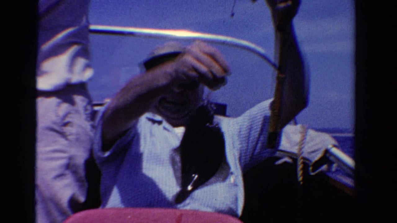 1958 Ontario home-movie frame with a seated adult man in a cap handling a fishing line while another person stands nearby on the boat.